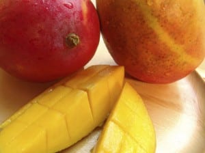 Two whole mangoes and a sliced mango on a wooden plate