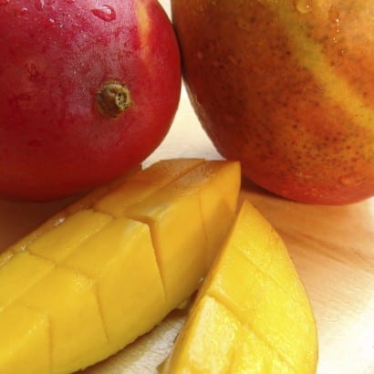 Two whole mangoes and a sliced mango on a wooden plate